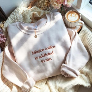 May include: A light beige sweatshirt with the embroidered text "Maybe coffee is addicted to me." A gold necklace with a circular pendant rests on the sweatshirt. A cup of coffee with latte art is in the background.