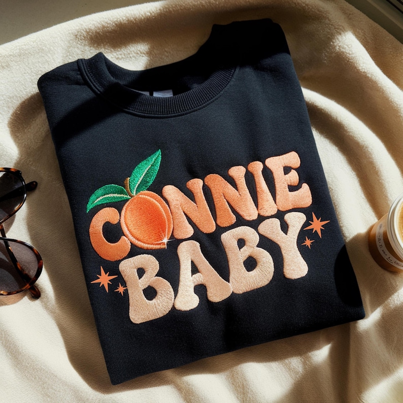May include: Black crew neck sweatshirt featuring the embroidered text "CONNIE BABY" in peach and cream, a peach graphic, and star details. Sunglasses and a candle are in the background.