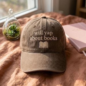 Peut inclure: Casquette de baseball marron avec le texte brodé "will yap about books" et un dessin de livre ouvert. Le chapeau est fait d'une matière douce et usée et a une visière incurvée. Un carnet rose et un terrarium en verre sont en arrière-plan.