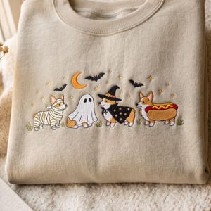 May include: A beige crewneck sweatshirt featuring an embroidered design of four Corgi dogs in Halloween costumes. The dogs are dressed as a mummy, a ghost, a witch with a hat and cape, and a hot dog, surrounded by a crescent moon, bats, and stars.