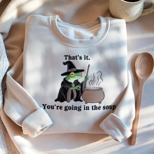 May include: Cream-colored sweatshirt featuring a cartoon frog dressed as a witch, stirring a cauldron. The text "That's it. You're going in the soup" is printed on the front. A wooden spoon and a mug are visible in the background.