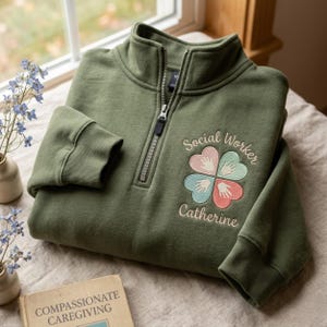 May include: Olive green quarter-zip sweatshirt with the embroidered text "Social Worker Catherine" and a four-leaf clover design with hand-shaped hearts in pink, blue, and peach. The sweatshirt has ribbed cuffs and a high collar.