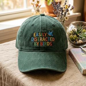 Könnte beinhalten: Dunkelgrüne Baseballkappe mit dem gestickten Text "EASILY DISTRACTED BY BIRDS" in bunten Buchstaben. Über dem Wort "BIRDS" ist ein blauer Vogel gestickt. Die Kappe hat einen gebogenen Schirm und einen Knopf oben.