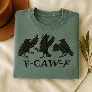 May include: A sage green crewneck sweatshirt featuring an embroidered design of three black crows in various poses. Below the birds, the text F-CAW-F is embroidered in black thread. The sweatshirt has a ribbed collar and long sleeves.