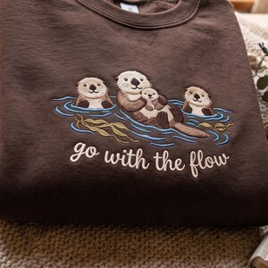 Puede incluir: Una sudadera de cuello redondo color marr&oacute;n oscuro con un bordado de tres nutrias marinas flotando en agua azul con algas verdes. El texto "go with the flow" est&aacute; bordado en una tipograf&iacute;a cursiva de color crema bajo la ilustraci&oacute;n.