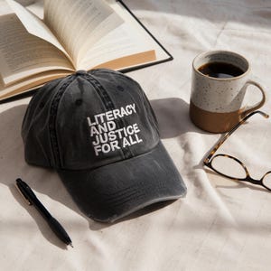 Puede incluir: Gorra de béisbol negra con el texto blanco "LITERACY AND JUSTICE FOR ALL". La gorra está colocada junto a una taza de café, un libro, un bolígrafo y gafas. La escena se desarrolla sobre una superficie blanca.