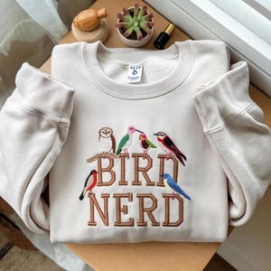 Op de afbeelding: Crèmekleurige sweatshirt met geborduurde vogels op een tak en de tekst "BIRD NERD" in bruin garen. Het ontwerp toont een uil, een kolibrie en andere kleurrijke vogels. De sweatshirt heeft geribde manchetten en een ronde hals.
