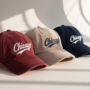 Könnte beinhalten: Drei Cord-Baseballkappen in Burgunderrot, Beige und Marineblau. Jede Kappe hat das Wort "Chicago" in einer stilisierten Schriftart gestickt. Die Kappen sind in einer Reihe angeordnet und zeigen ihre verschiedenen Farben und Texturen.