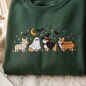 May include: A dark green crewneck sweatshirt featuring an embroidered design of four Corgi dogs in Halloween costumes. The dogs are dressed as a mummy, a ghost, a witch with a hat and cape, and a hot dog, surrounded by stars, a moon, and bats.