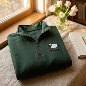 Könnte beinhalten: Ein dunkelgrünes Sweatshirt mit kurzem Reißverschluss und einer Stickerei auf der linken Brust, die ein weißes Schaf mit schwarzem Gesicht und Beinen zeigt. Unter dem Schaf ist das Wort Found in schwarzem Garn gestickt. Metallreißverschluss.