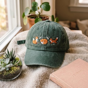 May include: A dark green baseball cap featuring an embroidered design of three orange foxes in various poses, including one sitting, one curled up sleeping, and one running, accented with small white flowers and green leaf sprigs.