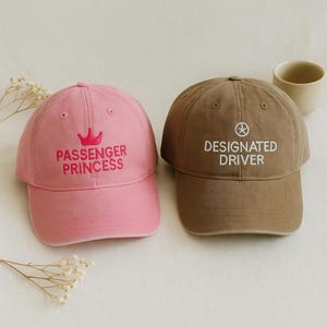 Pode incluir: Duas bonés de beisebol, um rosa e outro castanho. O boné rosa tem o texto "PASSENGER PRINCESS" bordado em rosa com um gráfico de coroa. O boné castanho tem o texto "DESIGNATED DRIVER" bordado em branco com um gráfico de volante.