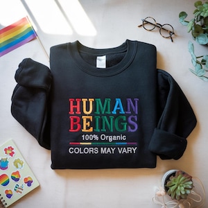 May include: Black sweatshirt with the embroidered text "HUMAN BEINGS" in rainbow colors. Below the text is "100% Organic" and "COLORS MAY VARY". A rainbow flag and stickers are visible.