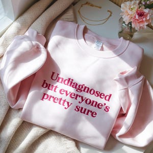 Könnte beinhalten: Ein hellrosa Sweatshirt mit dem Text "Undiagnosed but everyone's pretty sure" in dunklerem Rosa. Das Sweatshirt hat lange Ärmel und einen Rundhalsausschnitt. Das Bild ist aus der Vogelperspektive aufgenommen.