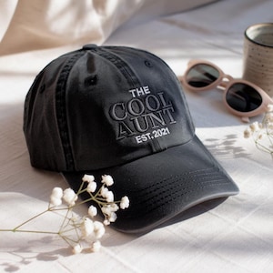May include: A dark grey baseball cap with the embroidered text "THE COOL AUNT EST. 2021". The cap has a curved peak and is made of a soft fabric. A pair of pink sunglasses and a ceramic mug are in the background.