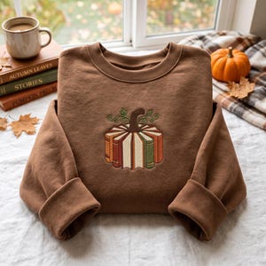 Pode incluir: Uma camisola castanha de gola redonda com um design bordado de uma abóbora construída a partir de livros verticais. Os livros estão dispostos num padrão circular com um caule castanho escuro e vinhas verdes no topo, centrados no peito.