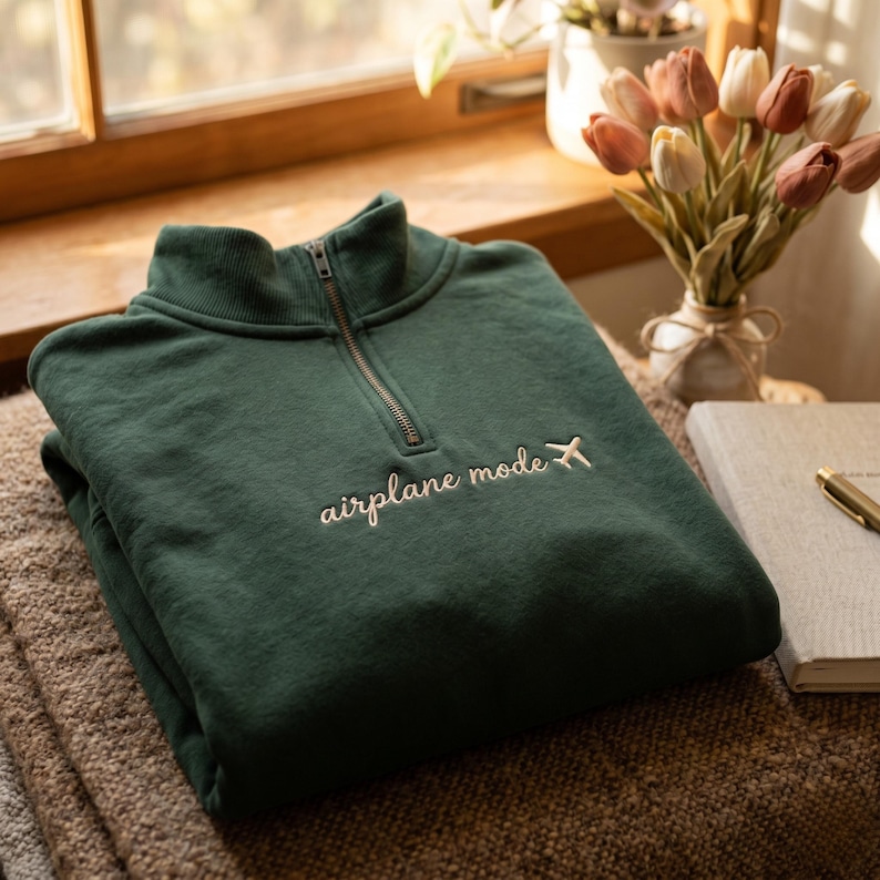 Puede incluir: Una sudadera verde oscuro con cuello alzado y cremallera de un cuarto. El texto "airplane mode" est&aacute; bordado en cursiva, acompa&ntilde;ado de un peque&ntilde;o gr&aacute;fico de avi&oacute;n. La sudadera est&aacute; doblada sobre una superficie texturizada.