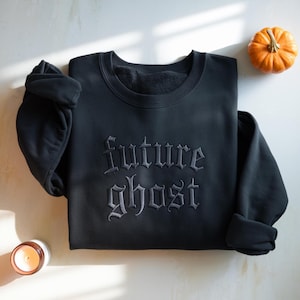 May include: A black long-sleeve crewneck sweatshirt featuring the words "future ghost" embroidered in a dark gray gothic-style font across the center of the chest. The garment has a ribbed neckline, cuffs, and hem with a soft interior lining.