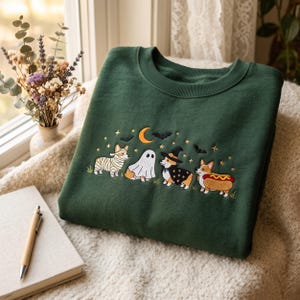 May include: A dark green crewneck sweatshirt featuring an embroidered design of four Corgi dogs in Halloween costumes, including a mummy, a ghost, a witch, and a hot dog, surrounded by a crescent moon, stars, and black bats.