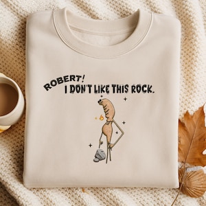 May include: Cream-colored sweatshirt with the embroidered text "ROBERT! I DON'T LIKE THIS ROCK." A cartoon figure with a flame and a rock is also embroidered on the front. The sweatshirt is folded on a patterned surface.