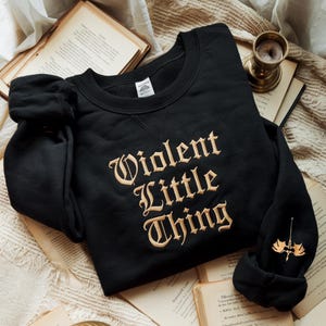May include: Black sweatshirt with the embroidered text "Violent Little Thing" in a vintage font. The sweatshirt has a crew neck and long sleeves. A small gold-colored emblem is on the left sleeve. The sweatshirt is displayed on a stack of old books.