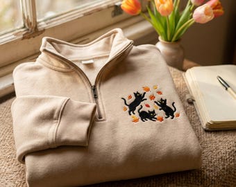 May include: A beige quarter-zip sweatshirt featuring an embroidered design of three black cats playing with scattered orange and yellow autumn leaves on the chest. The garment has a high collar with a metal zip and ribbed cuffs on the sleeves.