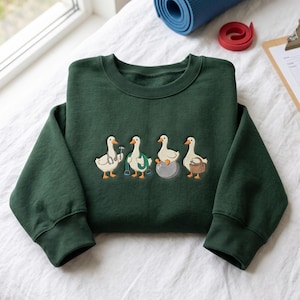May include: A dark forest green crewneck sweatshirt featuring an embroidered graphic of four white geese. The first goose wears a stethoscope, the second holds a jump rope, the third sits on a gray exercise ball, and the fourth carries a brown basket.