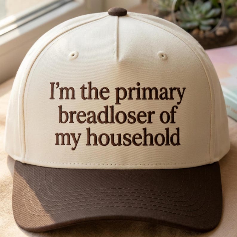 K&ouml;nnte beinhalten: Baseballkappe mit cremefarbener Krone und braunem Schirm. Auf der Kappe ist der Text "I'm the primary breadloser of my household" in braunen Buchstaben gestickt. Oben befindet sich ein brauner Knopf.