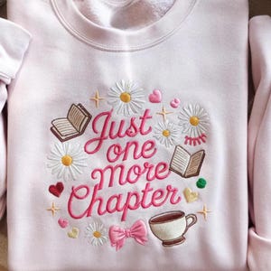 May include: A light pink sweatshirt featuring embroidered text that reads Just one more Chapter in a pink cursive font. The design includes white daisies with yellow centers, open books, a teacup, small hearts, a pink bow, and gold stars.