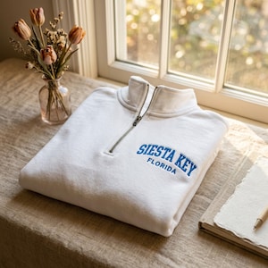 May include: A white quarter-zip sweatshirt featuring blue embroidered text on the chest that reads Siesta Key Florida. The garment has a high collar with a silver-toned metal zipper closure and long sleeves, presented in a folded, clean arrangement.
