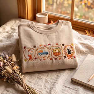 Peut inclure: Un sweat-shirt beige à col rond orné de broderies sur la poitrine représentant des motifs d'automne : une oie avec une écharpe, un camion bleu transportant des citrouilles, une citrouille aux tournesols et un chat noir dans une couronne.