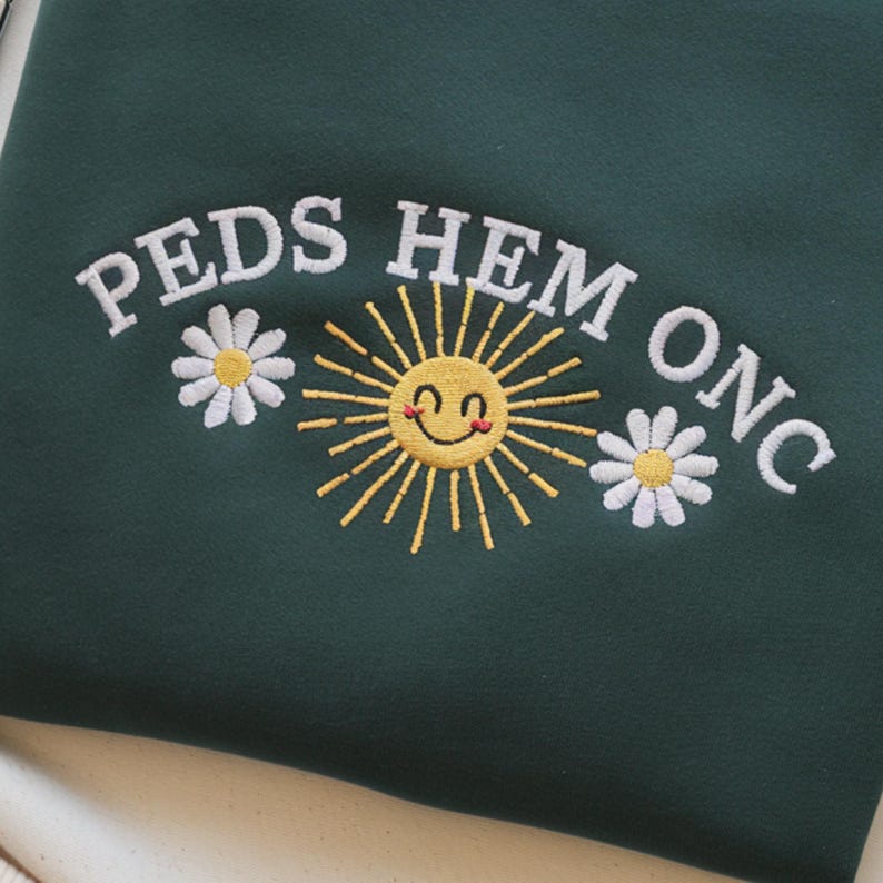 May include: Dark green sweatshirt with embroidered text "PEDS HEM ONC", a smiling yellow sun, and two white daisies. The design is centered on the front of the garment. The sweatshirt is made of a soft material.