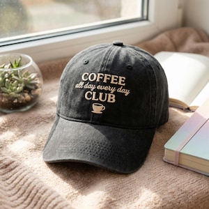May include: A black baseball cap with the words "COFFEE all day every day CLUB" embroidered in white. A small coffee cup graphic is below the text. The cap is sitting on a beige surface.