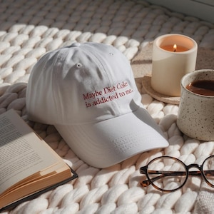 May include: White baseball cap with the embroidered text "Maybe Diet Coke is addicted to me." A lit candle, a mug of coffee, a pair of glasses, and an open book are also in the image. The cap is on a white, textured surface.