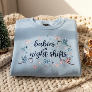 Puede incluir: Sudadera azul claro con el texto bordado "babies & night shifts". El diseño incluye ilustraciones de cigüeñas que llevan bebés, lazos, corazones y estrellas en rosa y azul. La sudadera está doblada sobre una manta de punto.
