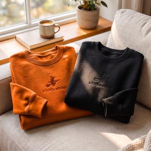 Könnte beinhalten: Zwei Rundhals-Sweatshirts, eines in gebranntem Orange und eines in Schwarz, jeweils mit einer Stickerei einer auf einem Besen reitenden Hexe über dem Text "a little witchy" mittig auf der Brust sowie gerippten Bündchen und Ausschnitten.