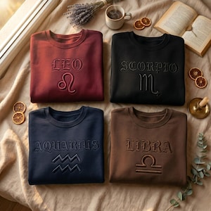 May include: Four crewneck sweatshirts in burgundy, black, navy, and brown, each embroidered with a zodiac sign and its name: Leo, Scorpio, Aquarius, and Libra. The sweatshirts are arranged on a beige surface with decorative items.