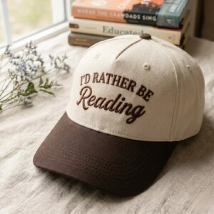 Peut inclure: Une casquette de baseball bicolore avec une calotte crème et une visière marron foncé. L'avant de la calotte présente le texte brodé I'D RATHER BE Reading en fil marron foncé. La casquette a une structure à cinq panneaux avec un bouton.