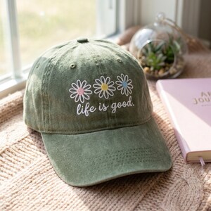 Op de afbeelding: Olijfgroene baseballcap met geborduurde madeliefjes in roze, geel en blauw. De tekst "life is good" is eronder geborduurd. De cap heeft een gebogen klep en een klassiek zes-panelen ontwerp.