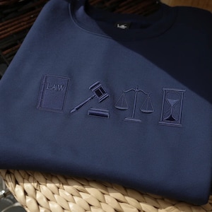 May include: Navy blue sweatshirt featuring embroidered law-related symbols: a book with the word "LAW", a gavel, scales of justice, and an hourglass. The symbols are in a darker shade of blue.