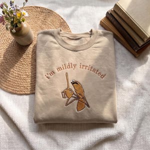 Può includere: Una felpa girocollo beige con testo ricamato in marrone che recita "i'm mildly irritated" sopra una formica ricamata in marrone che tiene un bastoncino, con un occhio mobile attaccato alla testa della creatura.