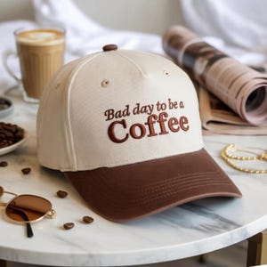 Puede incluir: Una gorra de béisbol de color crema con visera marrón oscuro y un botón en la parte superior. La parte frontal presenta un texto bordado en marrón oscuro que dice Bad day to be a Coffee, con la palabra Coffee en un tamaño de fuente mayor.