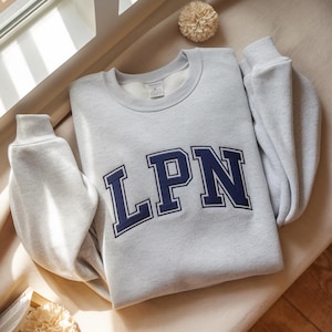 May include: A light grey sweatshirt featuring the navy blue letters "LPN" in a block font. The sweatshirt has a crew neck and long sleeves with ribbed cuffs. The letters are outlined in white. The garment is made of a soft material.