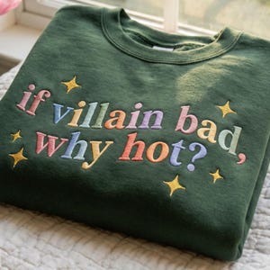 Op de afbeelding: Een donkergroene sweatshirt met de geborduurde zin "if villain bad, why hot?" in pastelkleuren. Gouden steraccenten omringen de tekst. De sweatshirt is netjes opgevouwen.