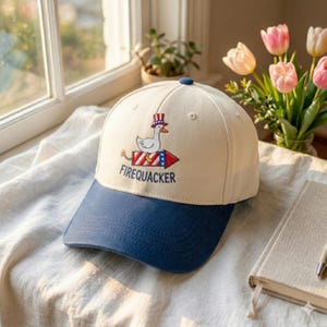 May include: A cream-colored baseball cap with a navy blue brim. The cap features an embroidered design of a duck wearing a top hat and a firework rocket with the text "FIREQUACKER".