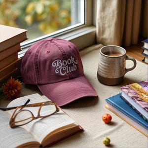Op de afbeelding: Een bordeauxrode baseballcap met de tekst "Book Club" in witte letters ligt naast een open boek en een bril. Een mok koffie en verschillende boeken staan ook op tafel. Een kleine oranje vrucht en een groene vrucht liggen ook op tafel.