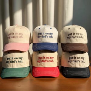 May include: A stack of baseball caps in various colors, including pink, navy, brown, red, green, and black. Each cap has a cream-colored crown and a colored brim. The phrase "put it on my my dad's tab." is embroidered on the front of each cap.