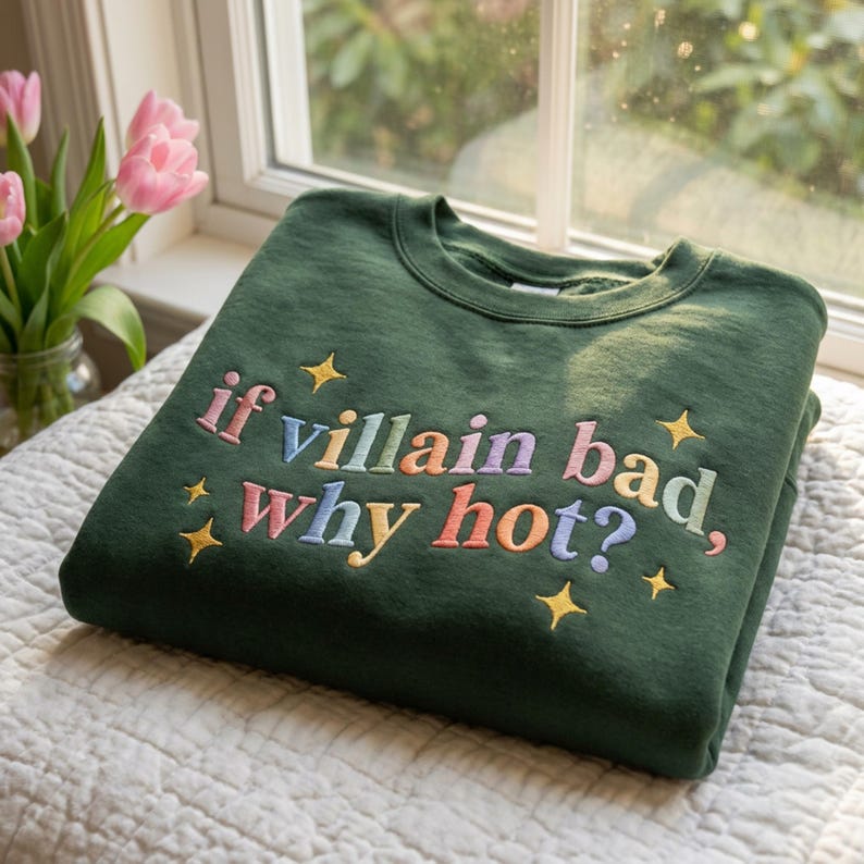 Op de afbeelding: Een donkergroene sweatshirt met de geborduurde zin "if villain bad, why hot?" in kleurrijke letters. Gouden steraccenten omringen de tekst. De sweatshirt is op een wit oppervlak gevouwen, met roze tulpen op de achtergrond.