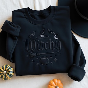 Könnte beinhalten: Schwarzes Sweatshirt mit dem Wort "Witchy" in gotischer Schrift gestickt, zusammen mit einem Hexenhut, Fledermäusen, einem Mond, einem Besen und einem Kessel. Das Sweatshirt ist von kleinen Kürbissen umgeben.
