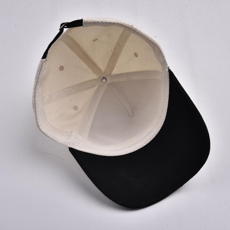 K&ouml;nnte beinhalten: Baseballkappe mit schwarzem Schirm und cremefarbener Krone. Die Innenseite ist cremefarben mit einem schwarzen, verstellbaren Riemen. Die Kappe wird von innen betrachtet und zeigt die N&auml;hte und die Konstruktion.